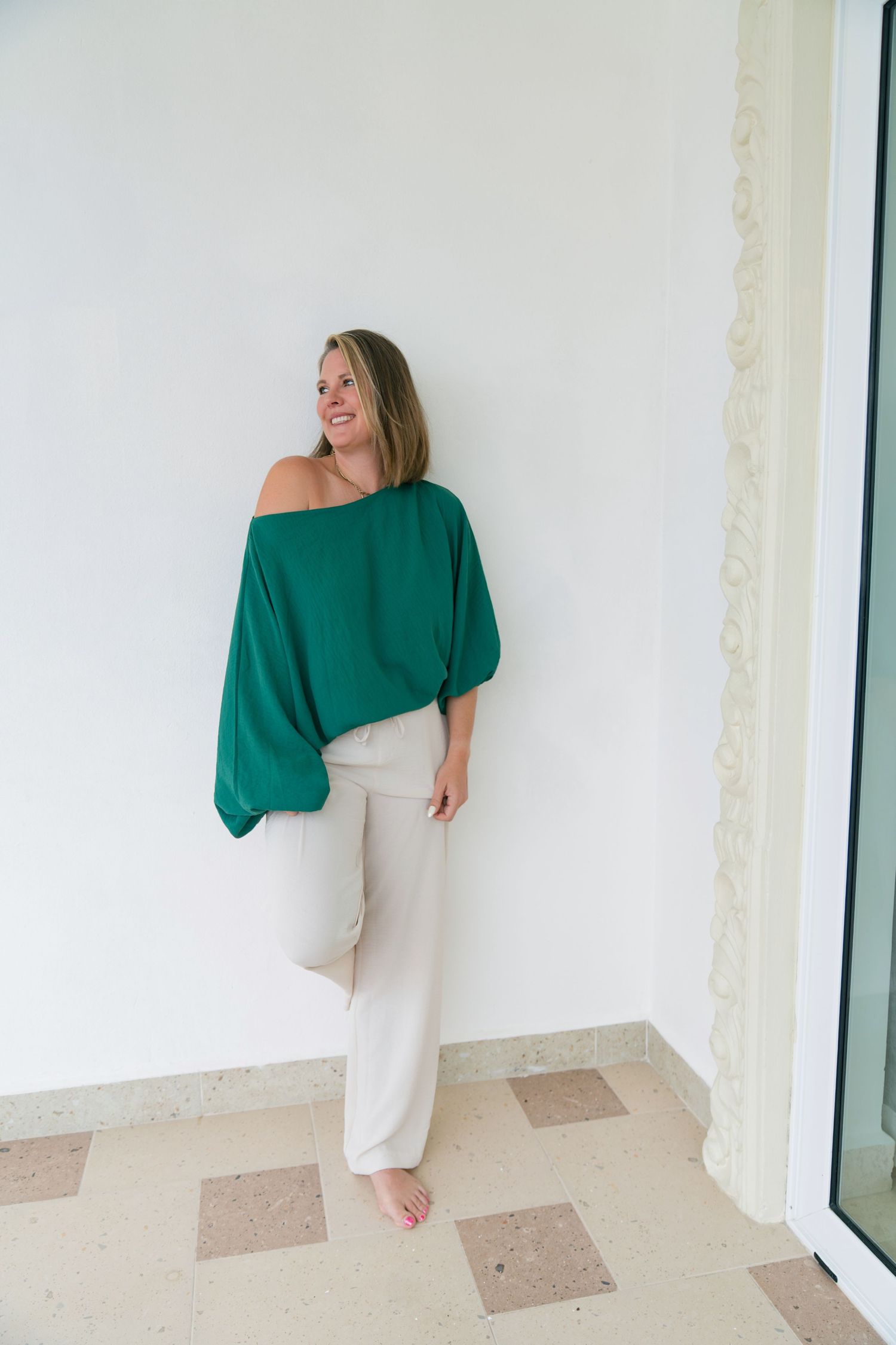 A person wearing a green off-shoulder sweater and white pants poses against a white wall in a casual indoor setting. Nebraska Photography