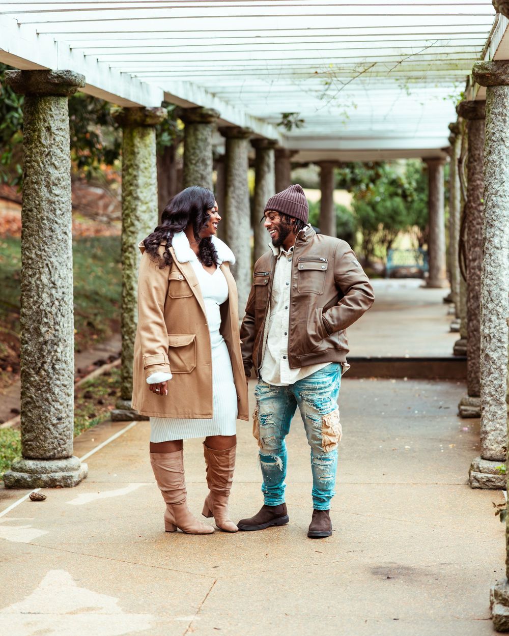 Couple sharing a smile while standing in columned garden walkway wearing coordinated fall outfits.