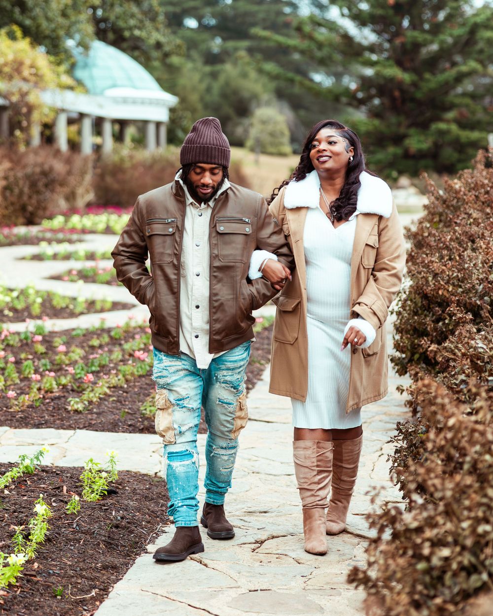 Couple strolling through formal garden path in autumn, woman in white dress and tan coat with knee-high boots.