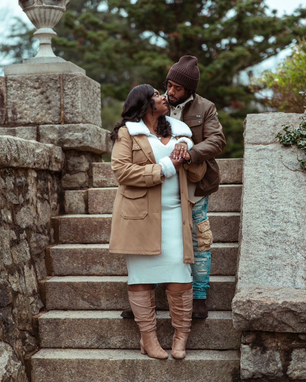 Couple sharing a kiss on stone steps outdoors, woman in white dress with tan coat and knee-high boots.