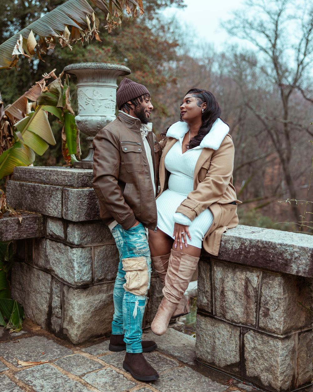Couple sharing a romantic moment on stone wall in garden, woman in white dress with tan coat and knee-high boots.