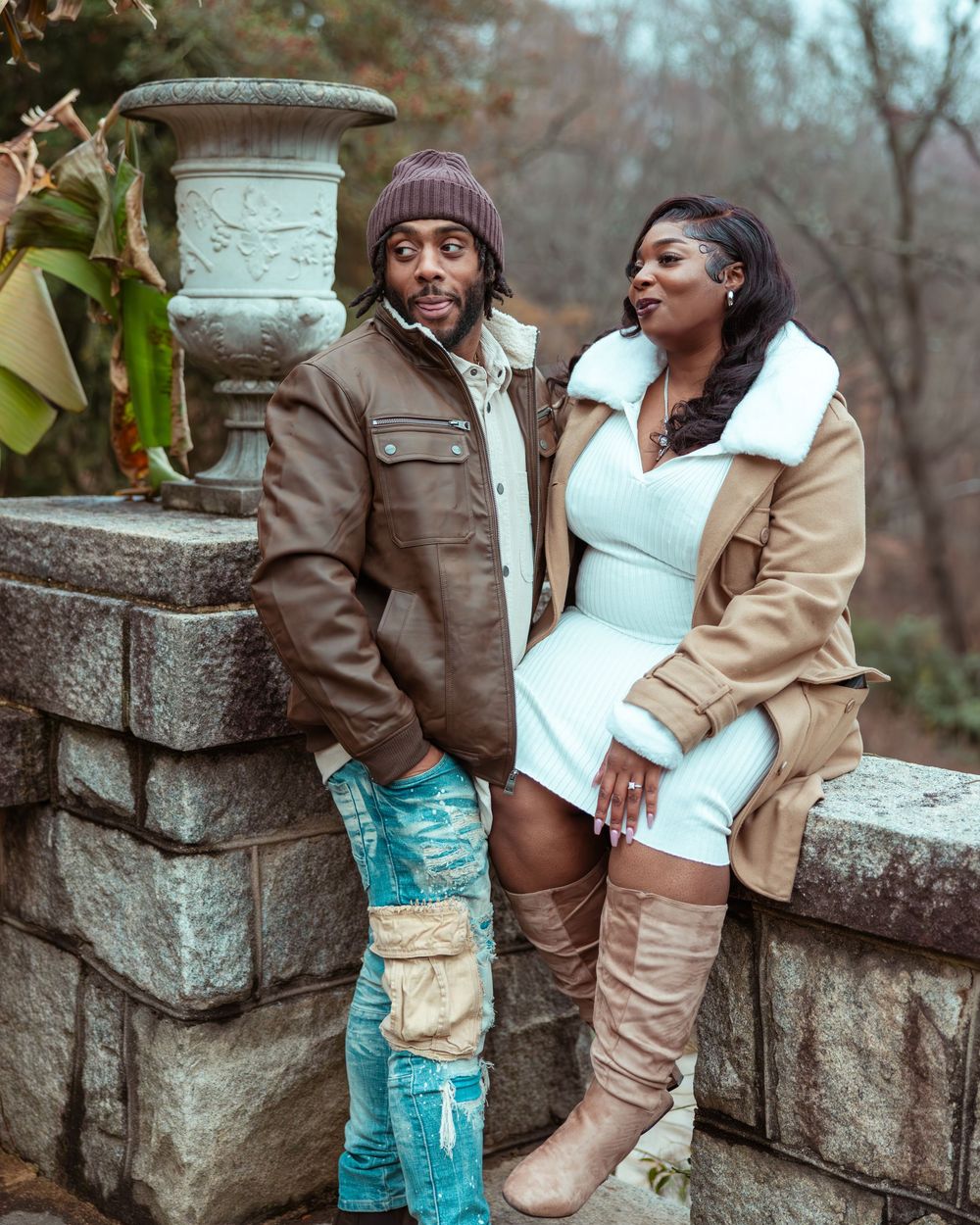 Stylish couple posing by stone wall in garden, man in distressed jeans and brown jacket, woman in white dress.