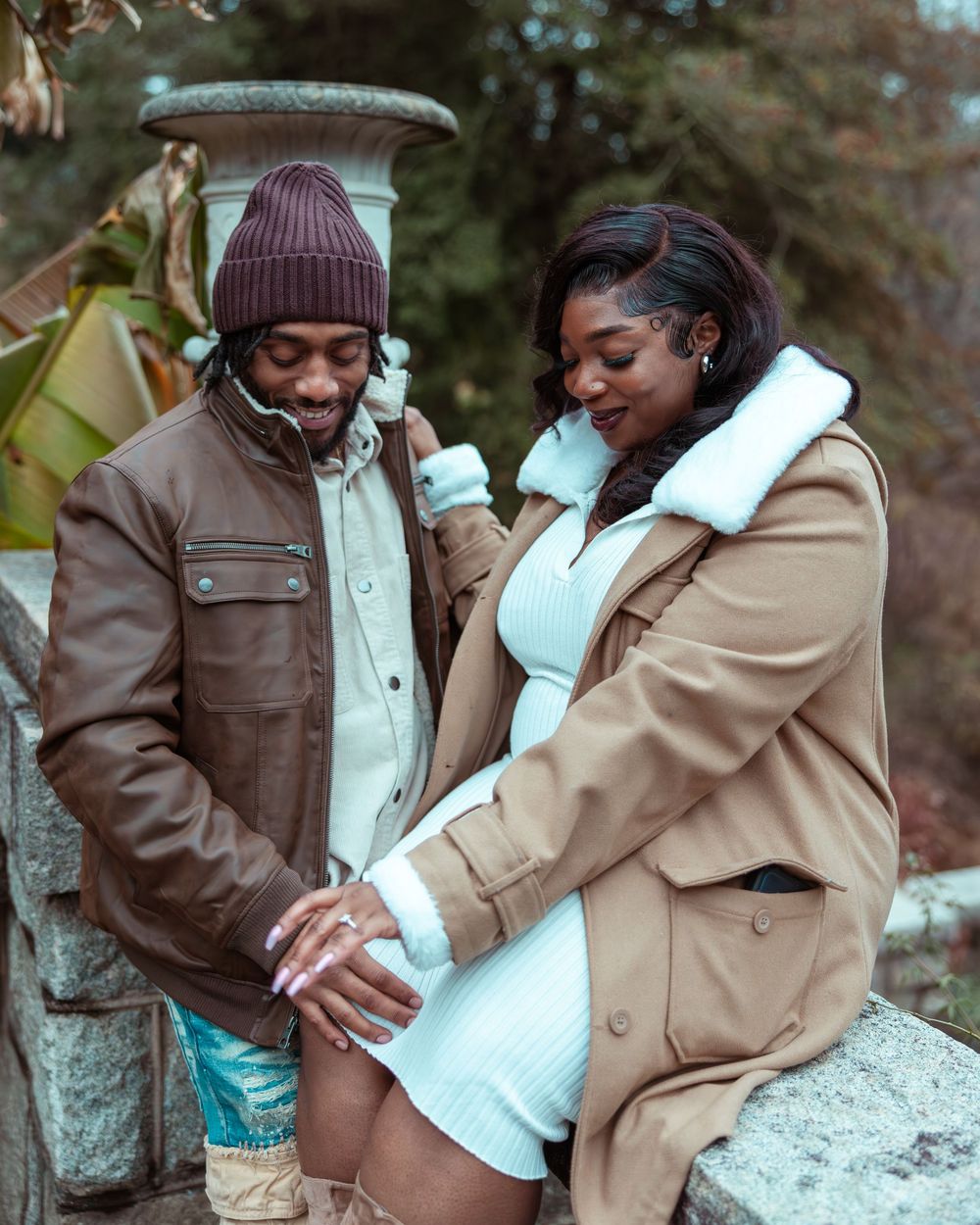 Man tenderly touching pregnant woman's belly outdoors, both wearing stylish fall outfits near stone wall.
