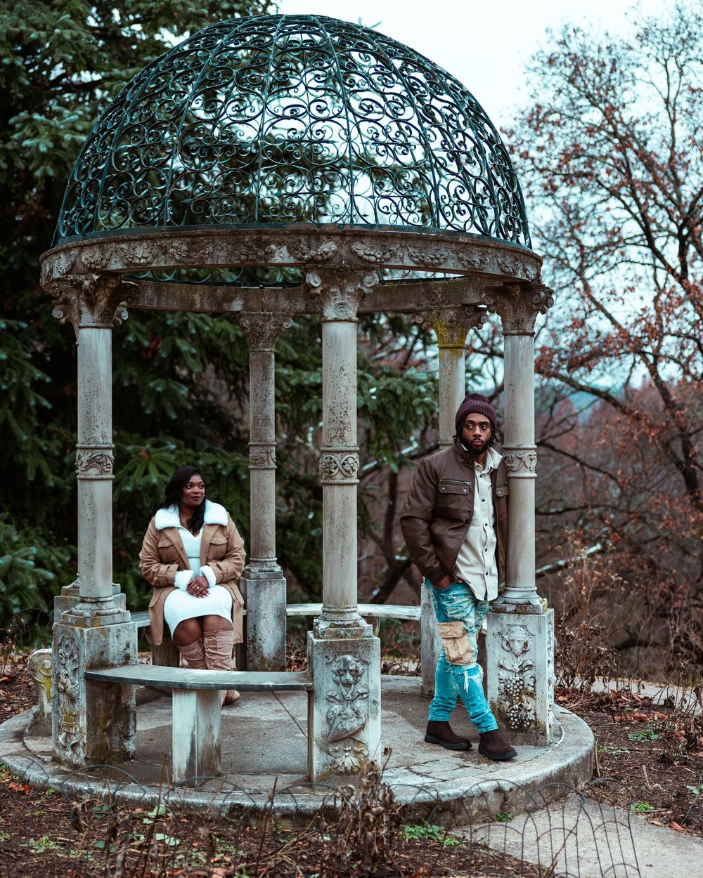 Engaged couple posing at an ornate stone gazebo surrounded by bare winter trees in a scenic garden setting.