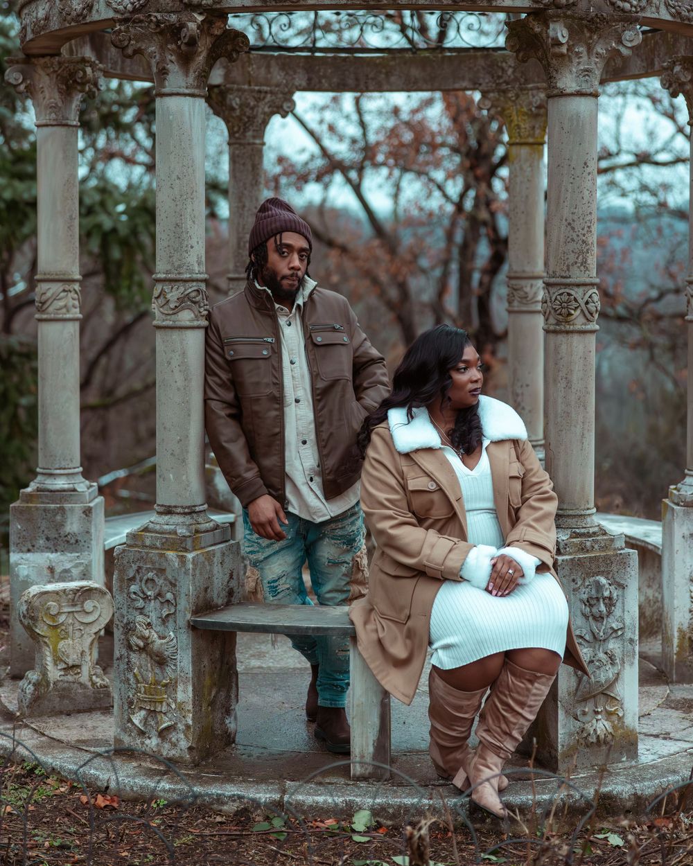 Couple posing under ornate stone gazebo in autumn park, woman in white dress and tan coat seated on bench.