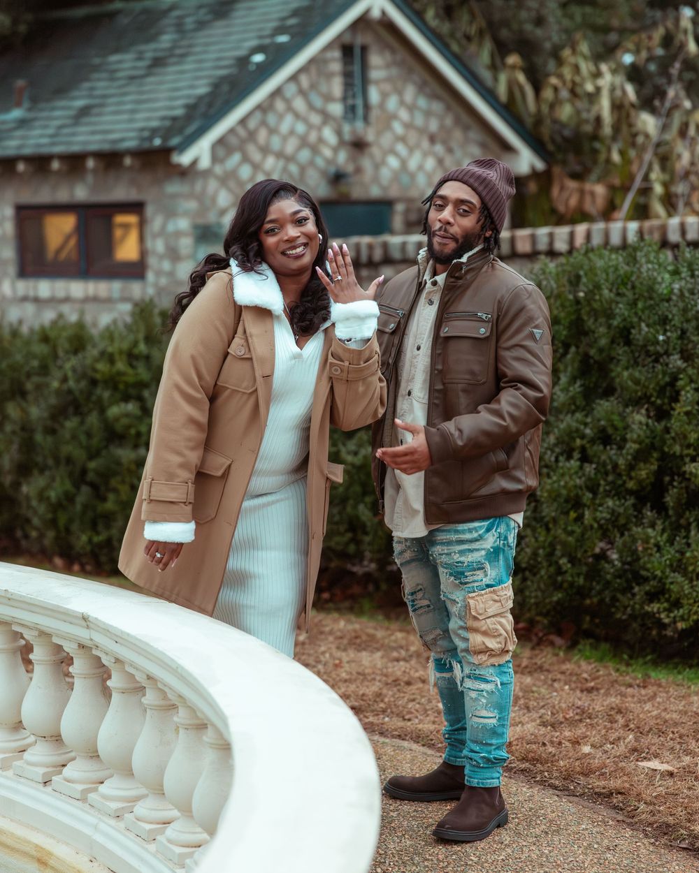 Newly engaged couple smiling outdoors near a stone estate, woman showing off her engagement ring to the camera.