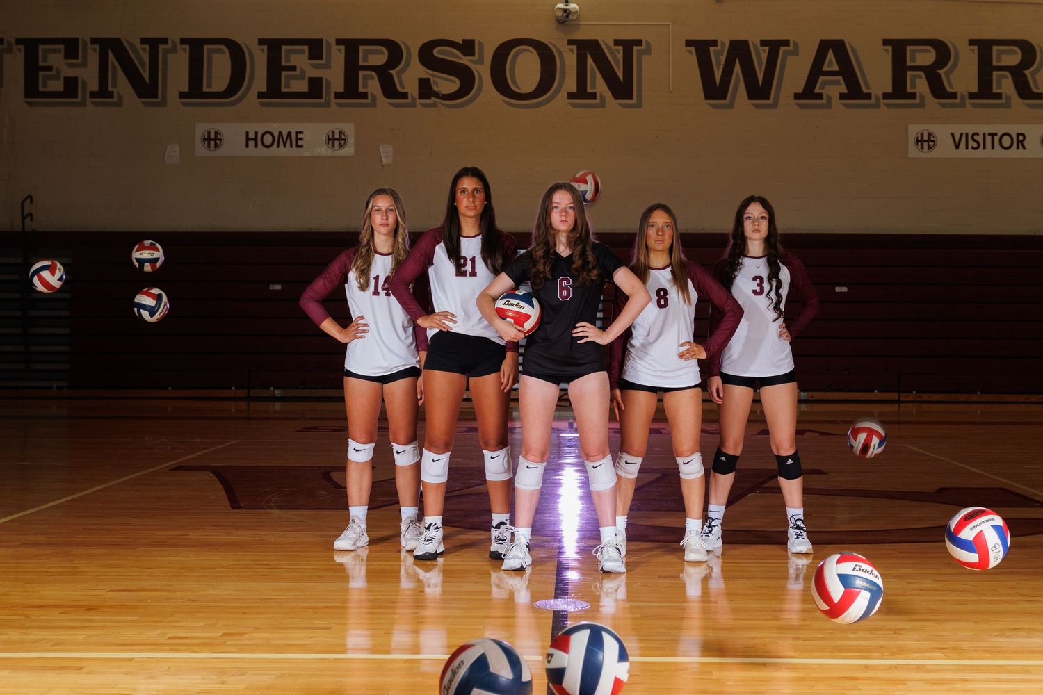Volleyball team members pose with scattered balls in Henderson Warriors gymnasium.