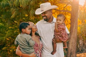 Smiling father in cowboy hat holding two young children under golden autumn trees.
