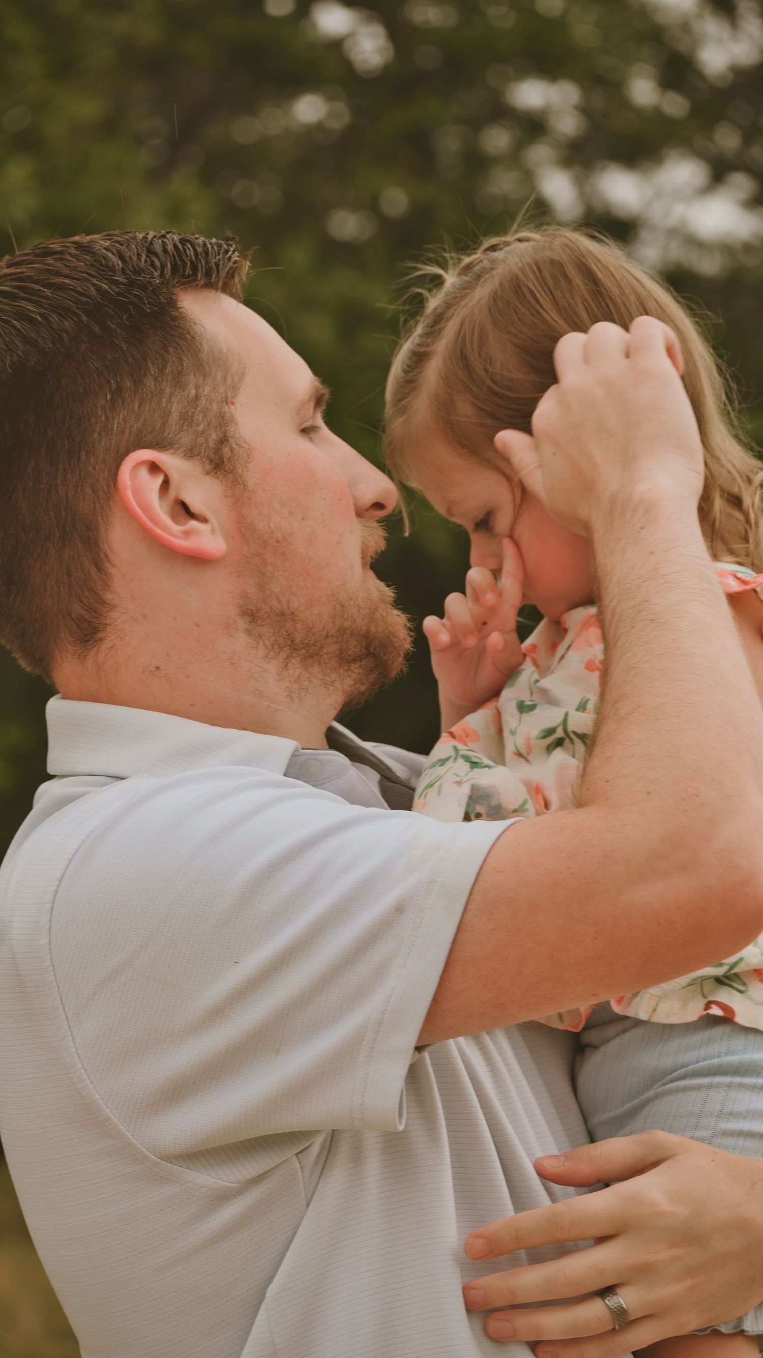 A father tenderly holds his young daughter close, gently touching her forehead in a warm outdoor setting.