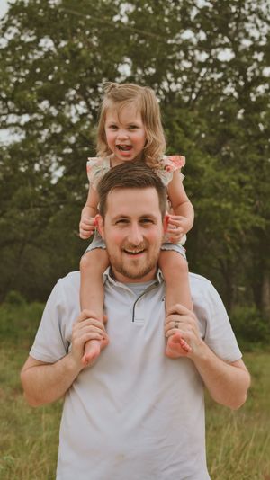 A smiling man carries a laughing toddler girl on his shoulders outdoors with green trees in the background.