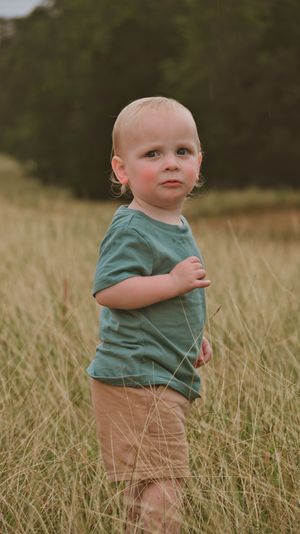 Toddler boy in green shirt and tan shorts standing in tall grass field.