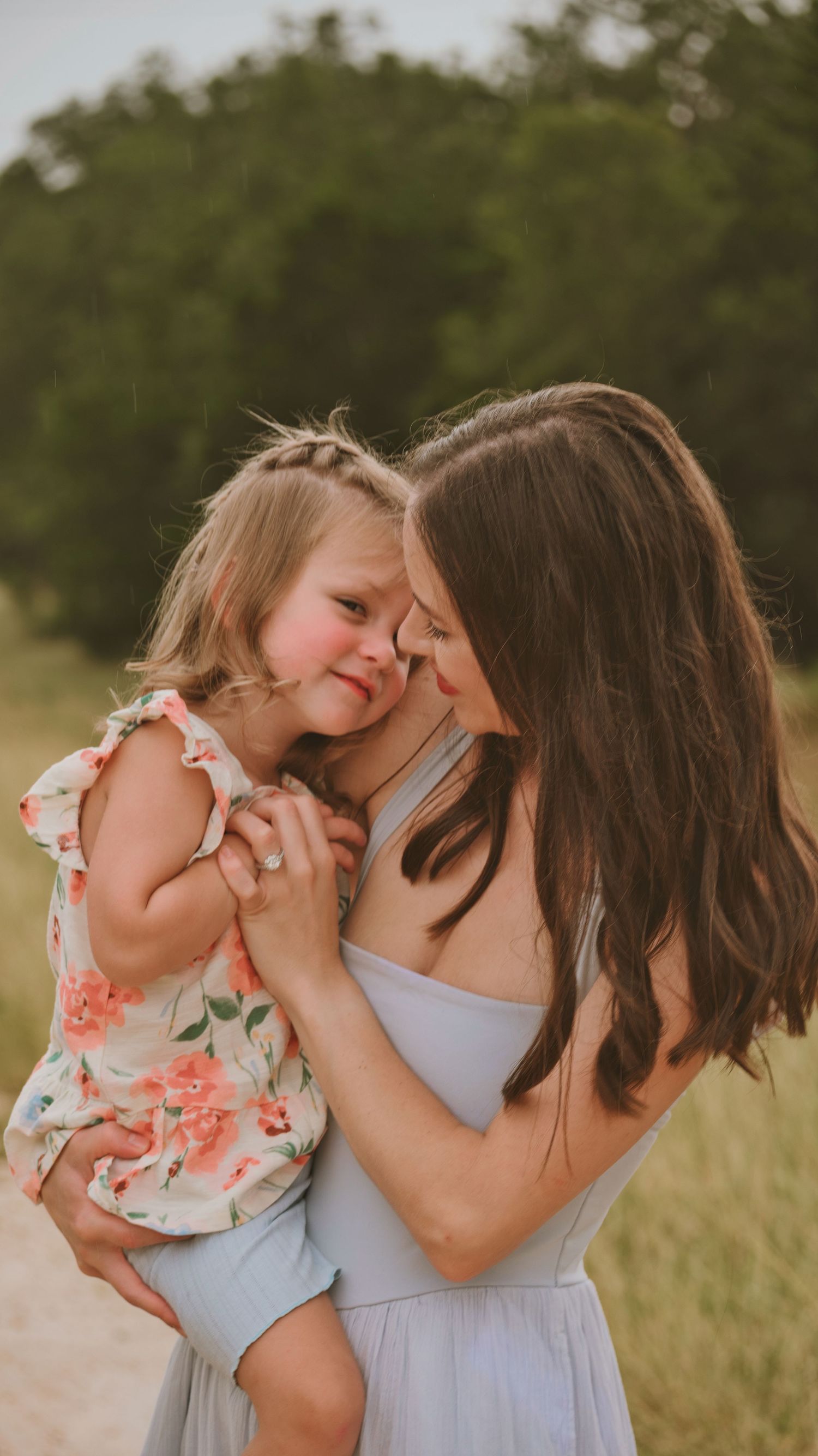 A mother tenderly holds her smiling toddler daughter in a floral dress outdoors in a golden field.