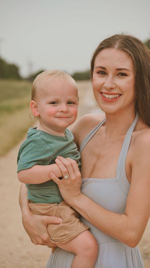 A smiling brunette mother holds her baby boy in a green shirt during an outdoor photo session.