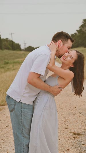 A couple embraces and shares a kiss on a rustic dirt path surrounded by green fields.