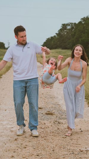 Happy family swinging laughing child between parents on rural dirt path.