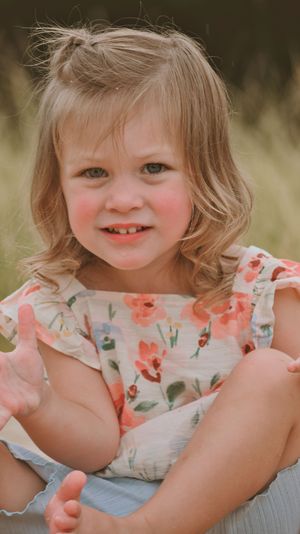 Smiling toddler girl with blonde hair wearing a floral dress, posed outdoors in a sunny field.