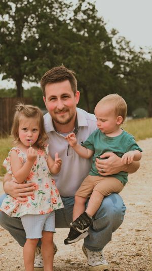 Father crouching with two young children on a dirt path, boy in teal shirt and girl in floral dress.