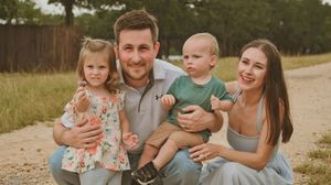 Happy young family of four posing outdoors on a dirt path, parents holding toddler girl and baby boy in warm sunlight.