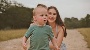 A young mother smiles warmly behind her toddler boy in a green shirt on a sunny dirt path surrounded by greenery.