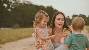 A mother crouches on a sandy path holding a flower while two young children interact with her outdoors.