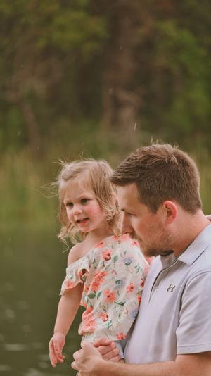 A father holds his young blonde daughter in a floral dress outdoors near a lake with green trees in the background.