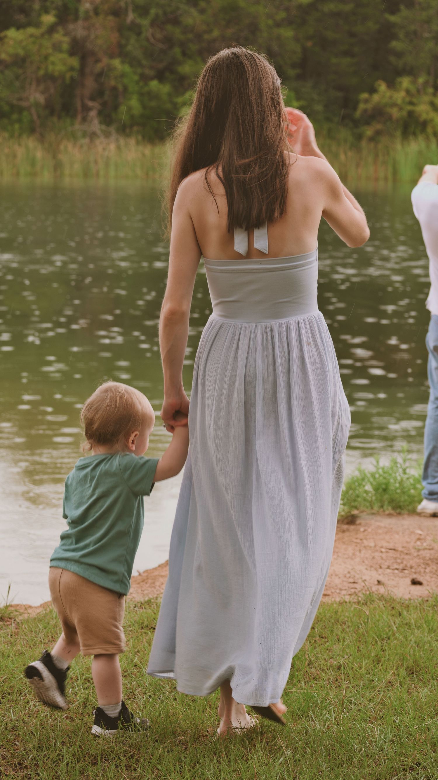 Mother in gray maxi dress holding toddler son's hand by peaceful lake.