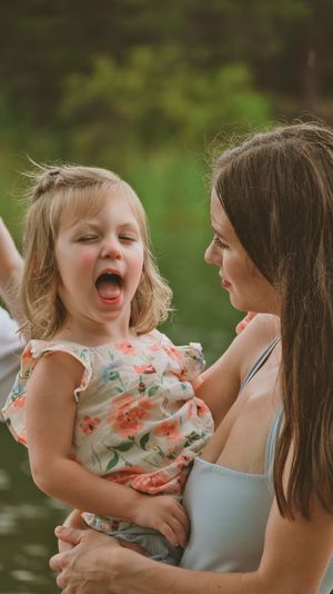 Mother in blue dress holds laughing toddler girl in floral dress outdoors with green trees in background.
