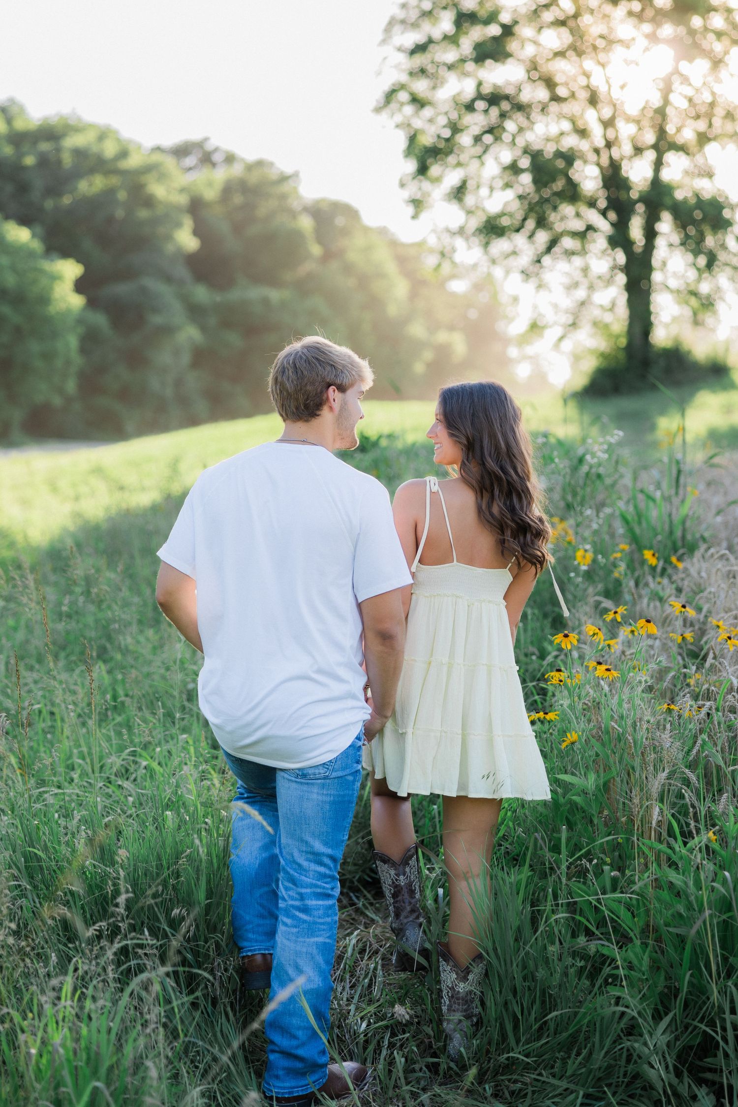 A couple walks together through a meadow at sunset holding hands.