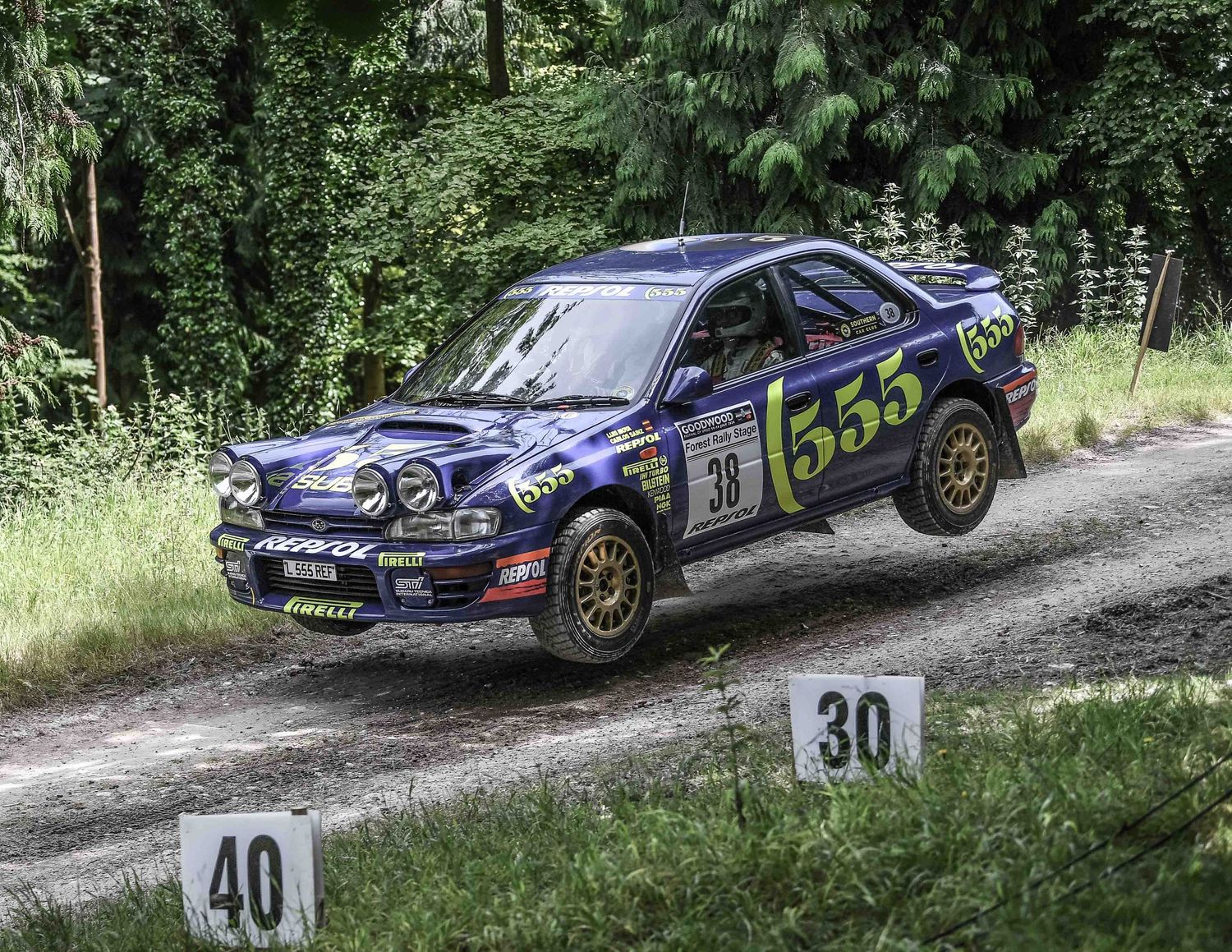 A blue Subaru WRX rally car navigates a dirt course through dense forest.