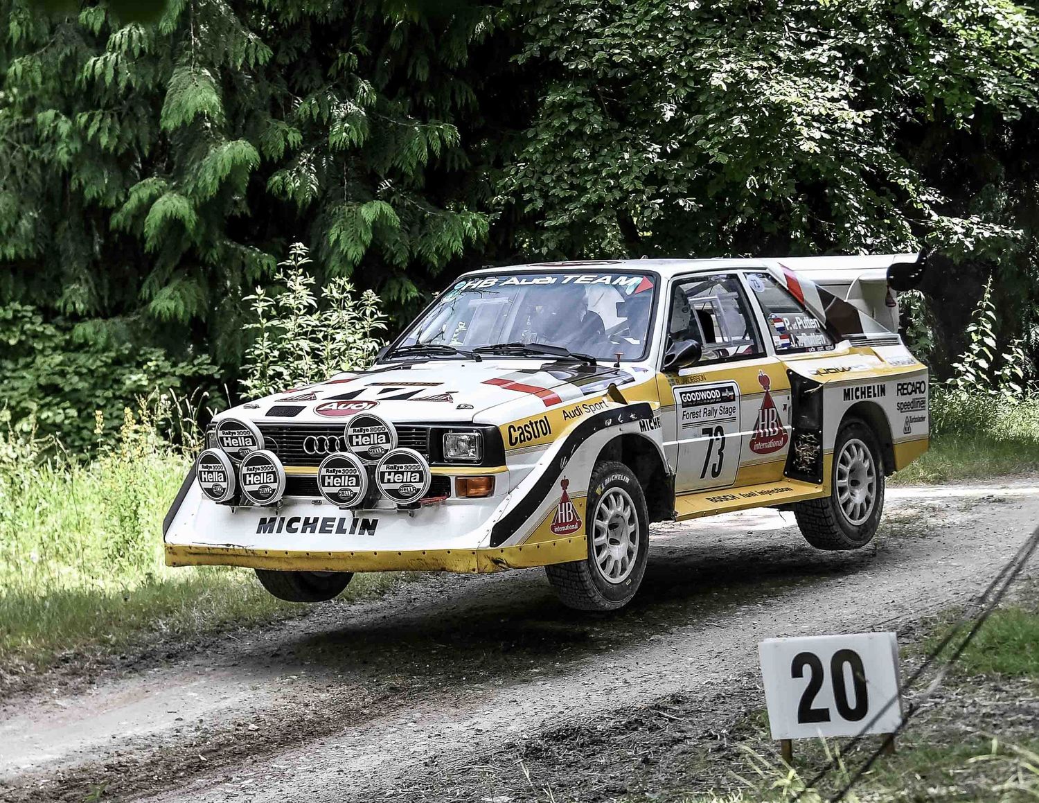 A white Audi Quattro rally car races through a forested dirt track marked with course numbers.