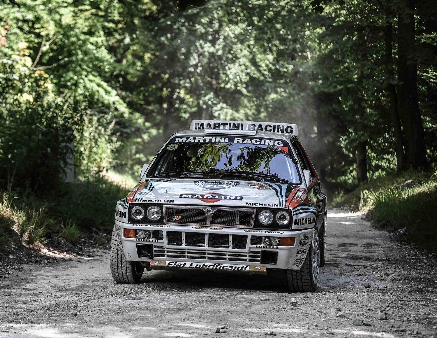 Lancia Delta Integrale rally car kicks up dust while racing through wooded course.