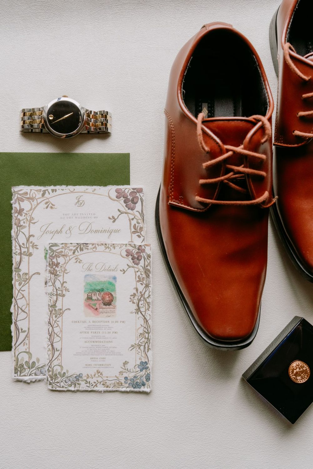 Wedding accessories including brown leather shoes, watch, and decorative invitation on white surface.