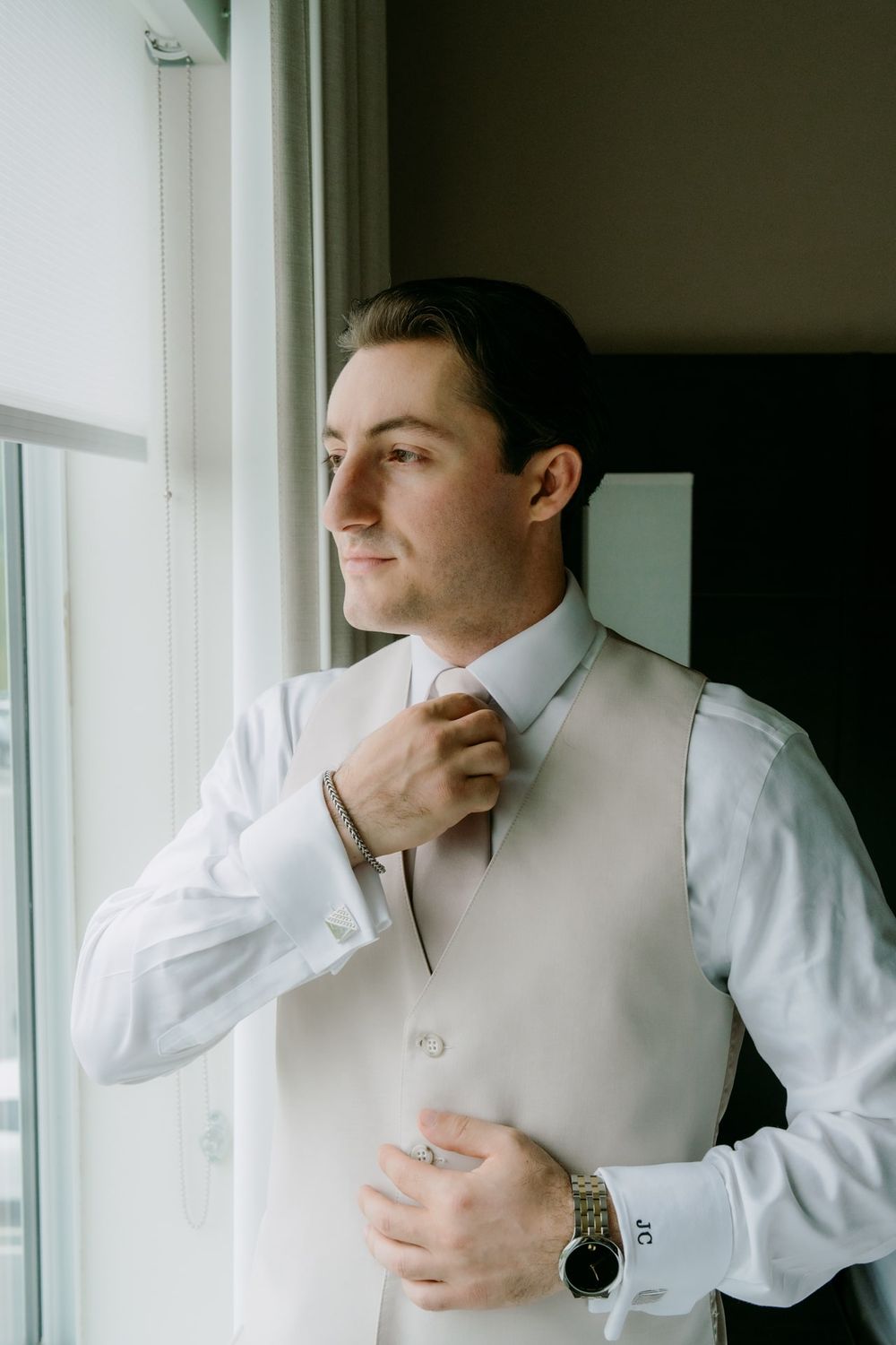 A person in formal attire adjusts their tie while standing by a window.