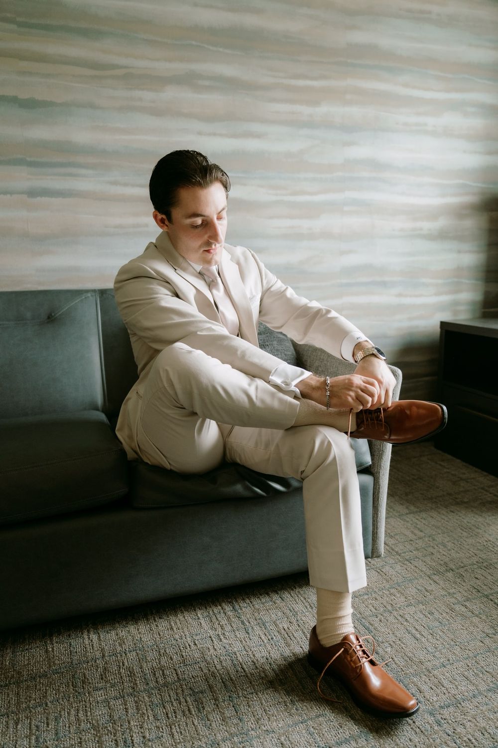 A person in a beige suit sits on a gray couch while adjusting their brown leather shoes.