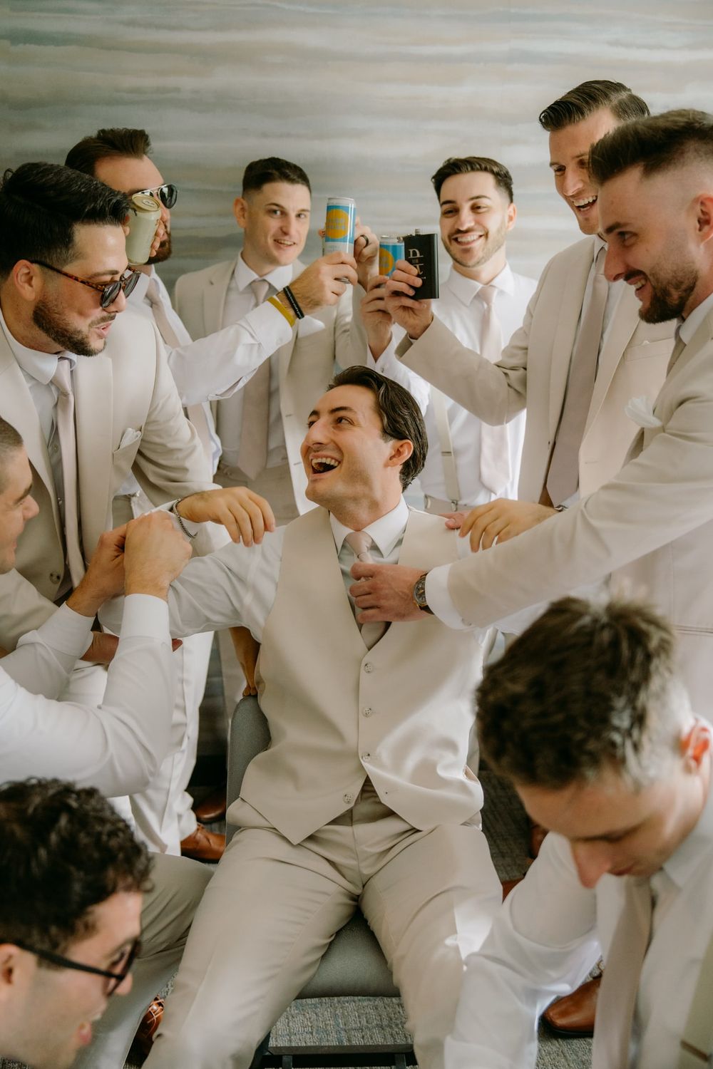 Groomsmen in light beige suits share drinks and laughter during a celebratory moment at a wedding.