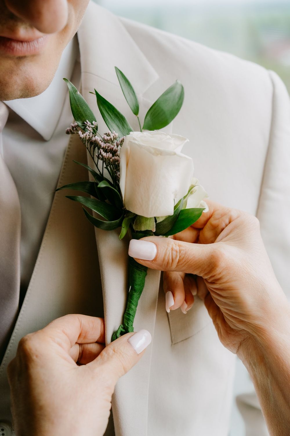 A white rose and greenery boutonniere being pinned onto a light beige suit lapel.