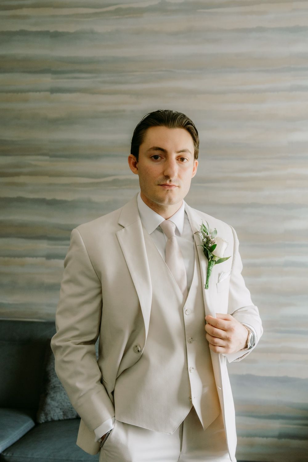 A groom wearing a light beige three-piece suit with a floral boutonniere poses against a gray backdrop.