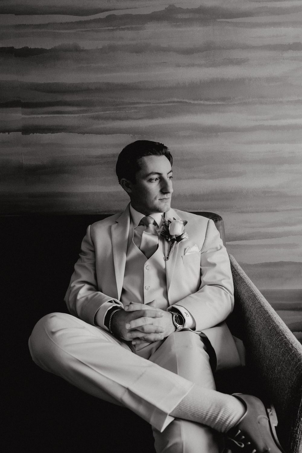 A black and white portrait of a groom in a light-colored suit seated against a textured backdrop.