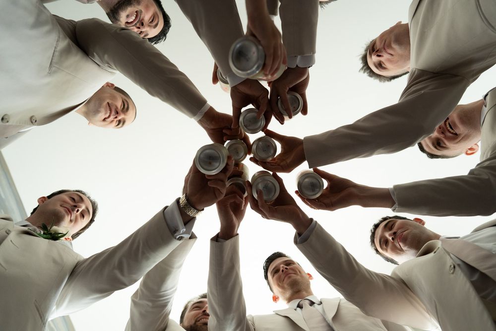 Groomsmen in white dress shirts form a circle to toast with drinks from above.