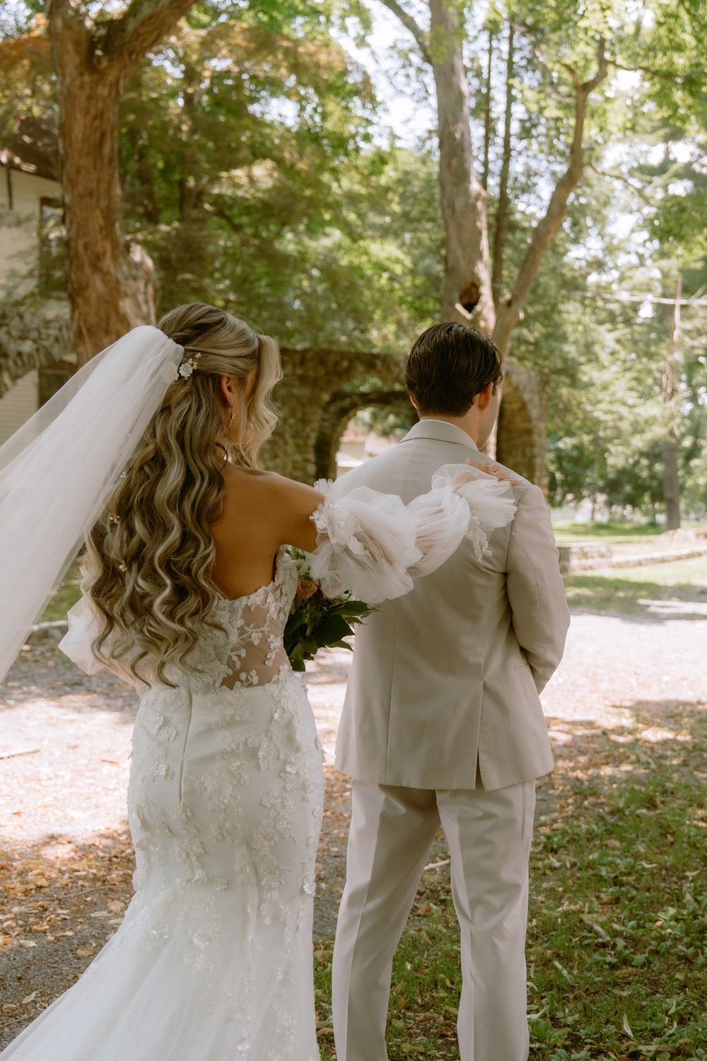 A bride in a lace wedding gown and long veil stands behind her groom in an outdoor setting.