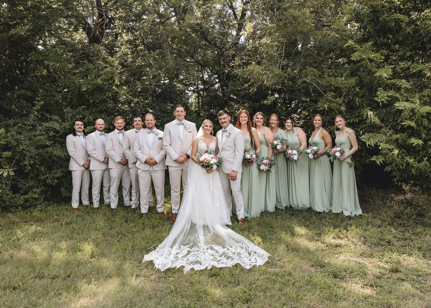 A wedding party wearing sage green and white dresses poses together on a grassy hillside in a natural outdoor setting. Neewton, KS