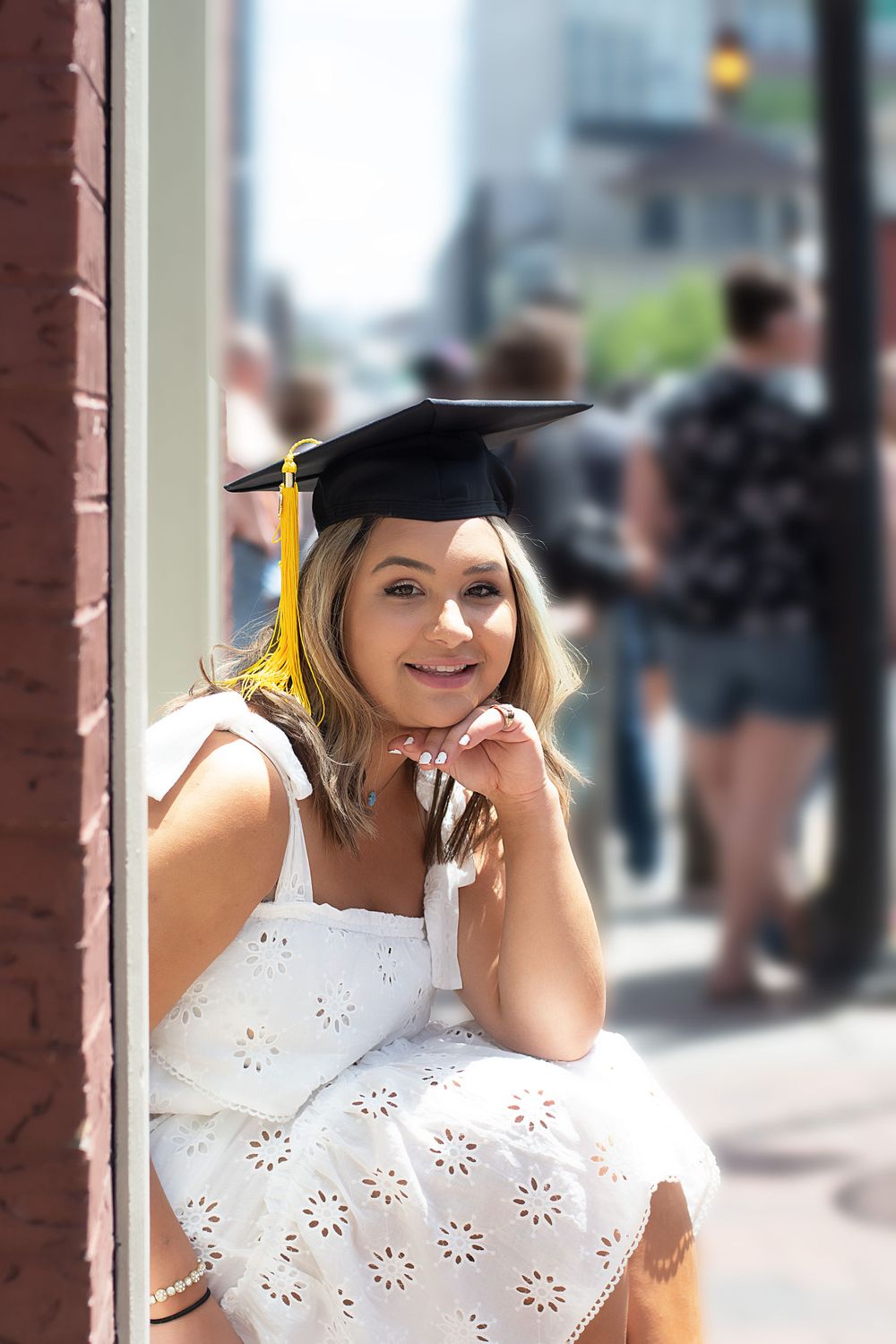 Graduate in white eyelet dress and cap poses outdoors near brick building.