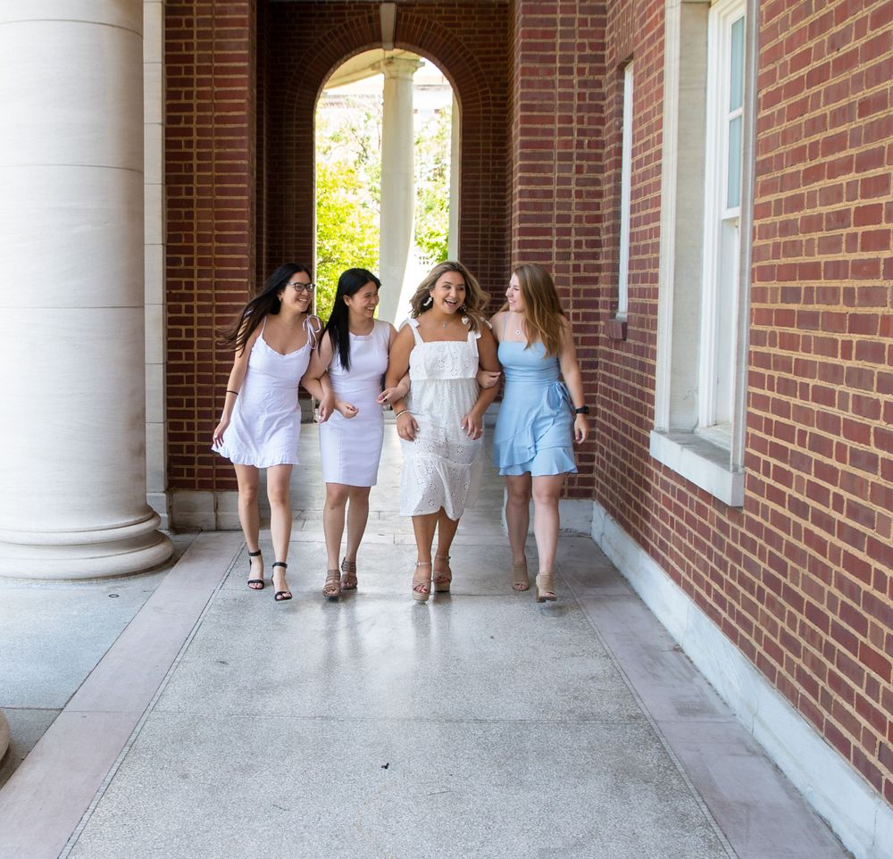 Four friends in white summer dresses walk together through columned university hallway.