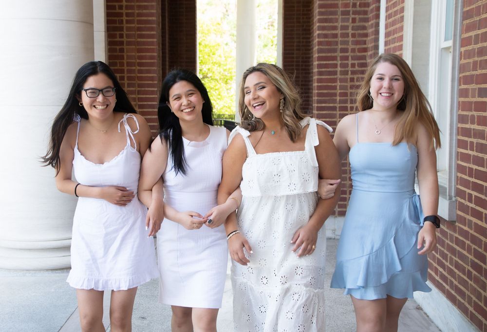 Four friends in light colored dresses smile together between white columns.