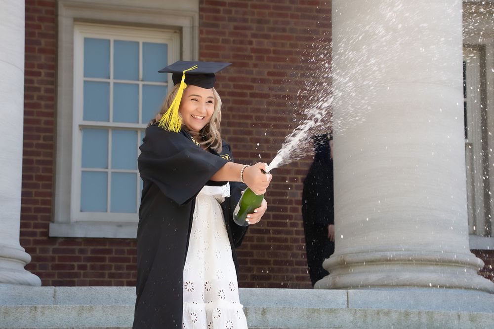 Graduate in cap and gown sprays champagne while celebrating on campus steps.