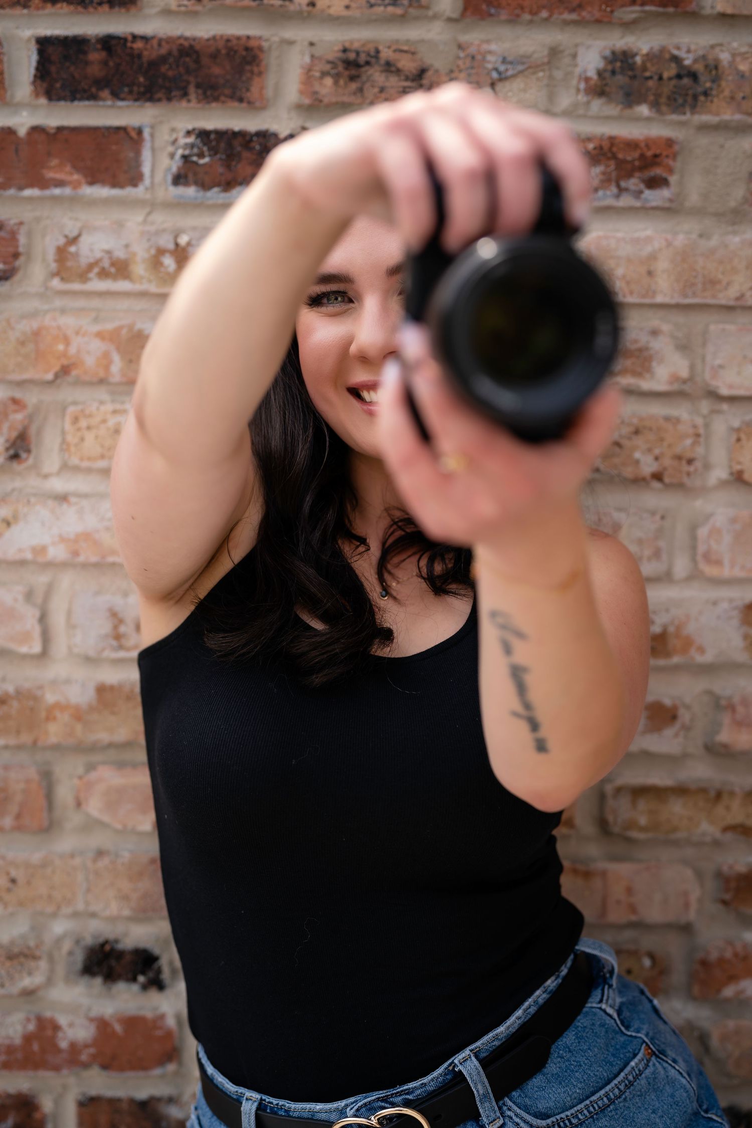 Professional photographer holding camera against brick wall wearing black tank top and jeans.