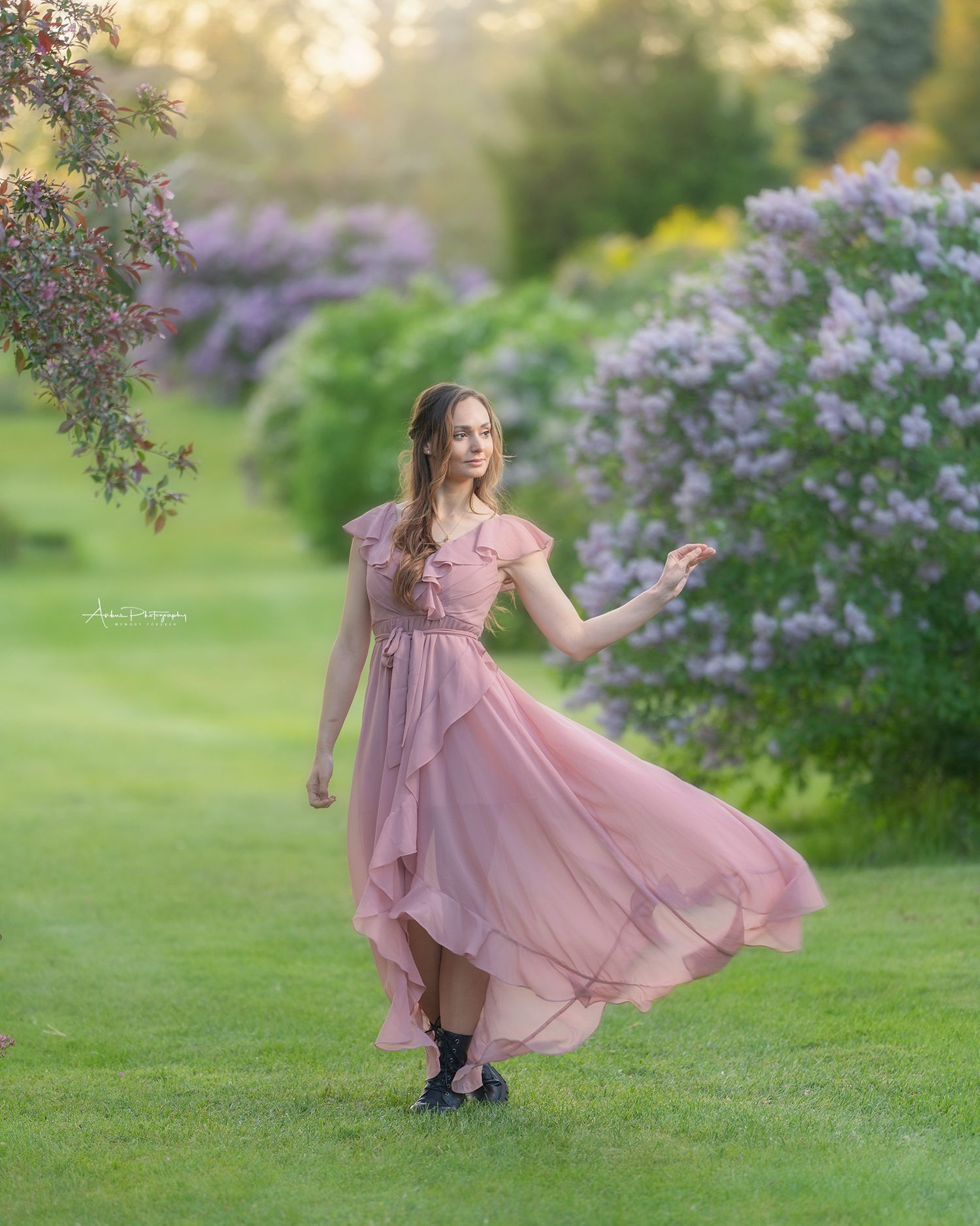 Ethereal pink dress flows in a garden with blooming lilac bushes during golden hour photography session.
