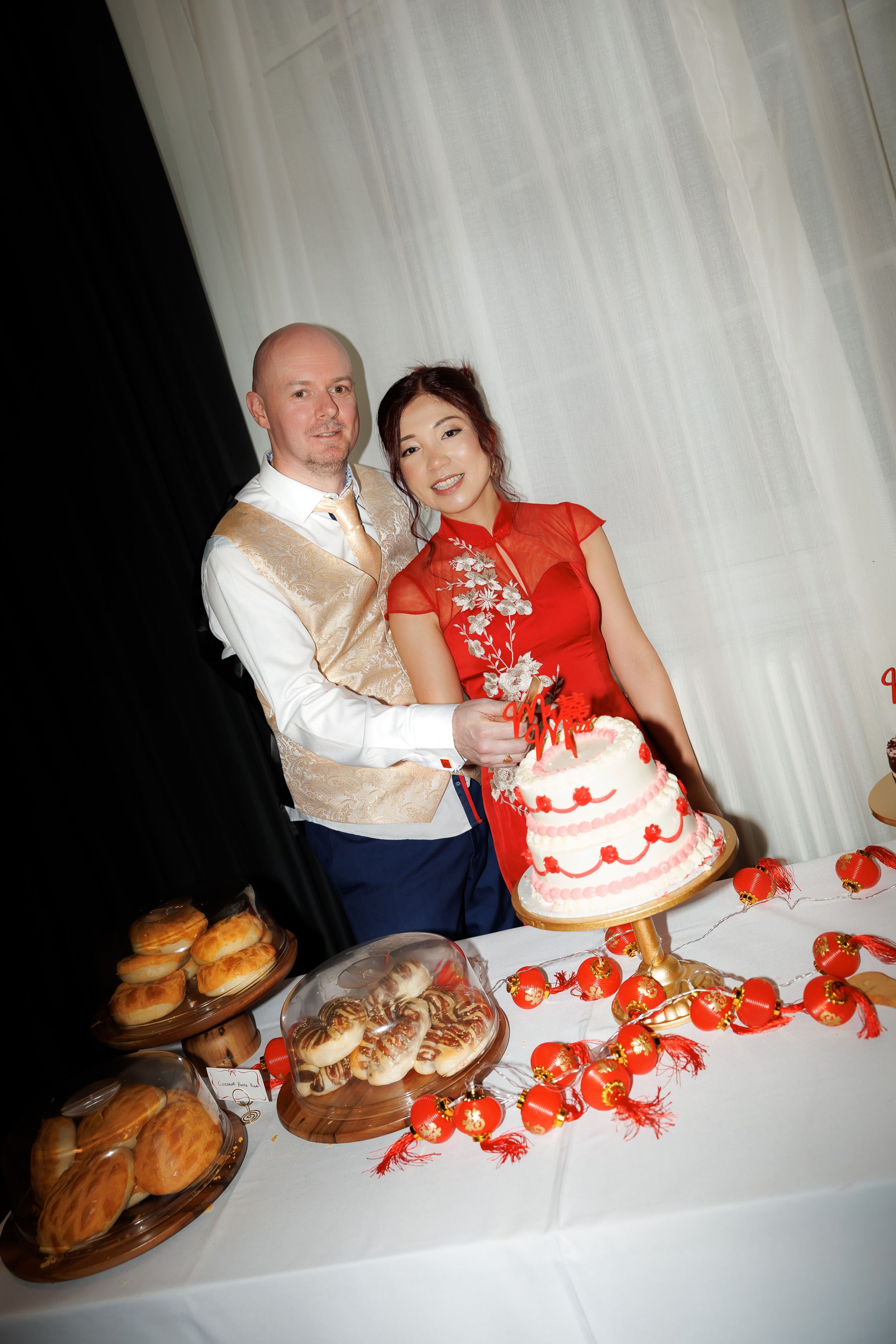 Couple in wedding attire cutting a white and red tiered cake at a dessert table with pastries and decorations.