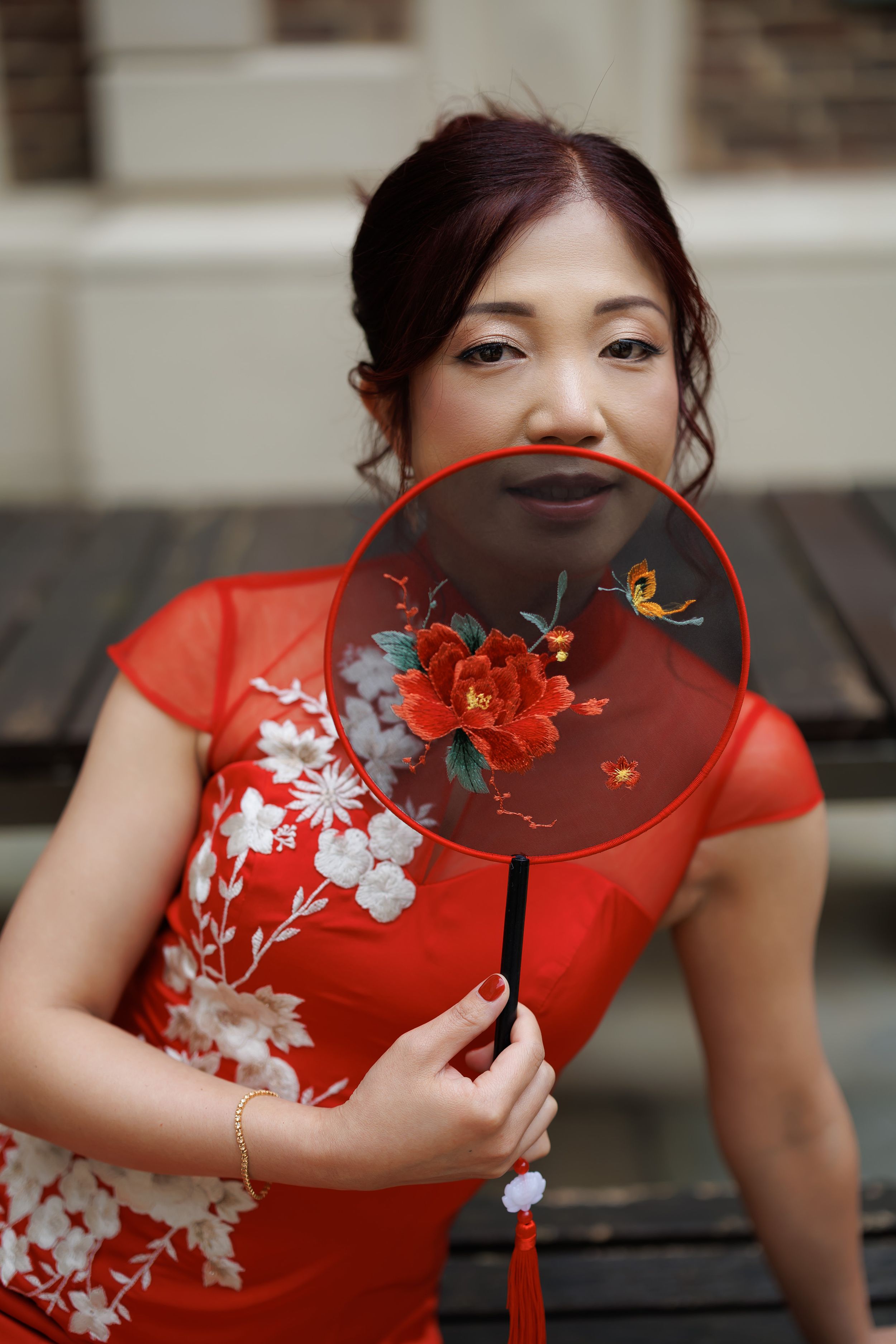 Woman in red floral cheongsam holding decorative red fan with peony embroidery, posing outdoors.