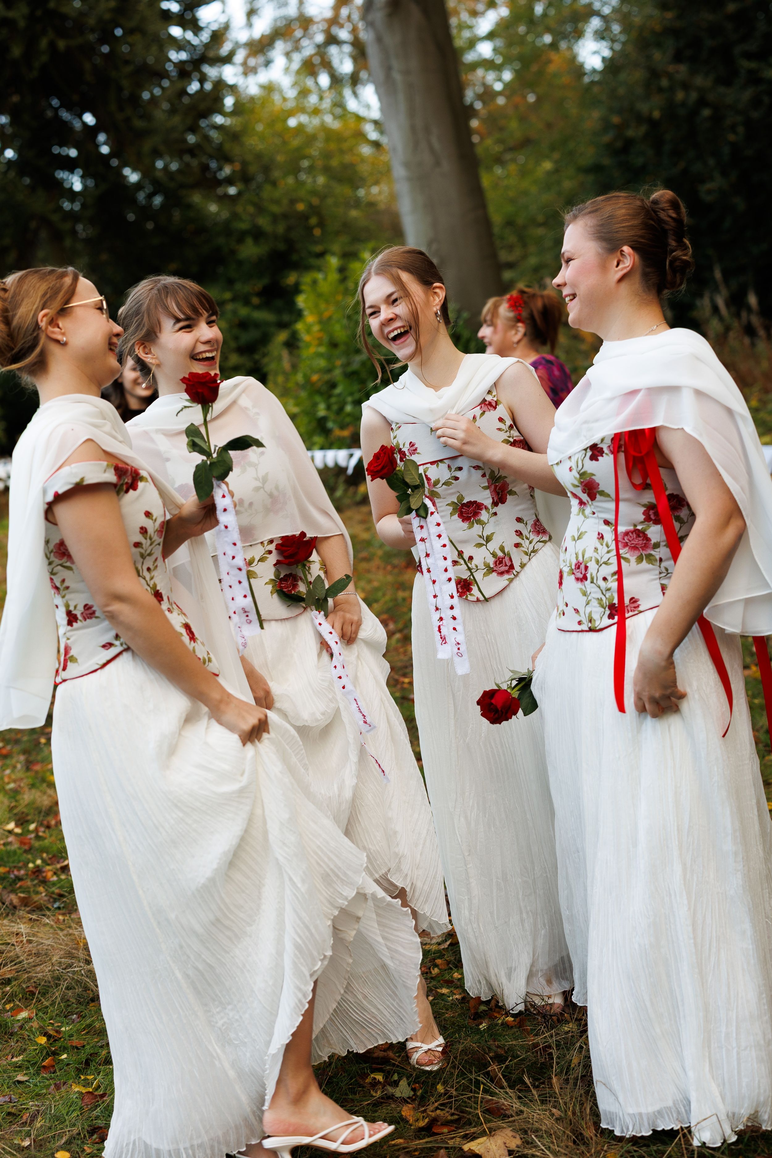Four young women in matching white floral dresses laughing and holding red roses outdoors at a garden event.